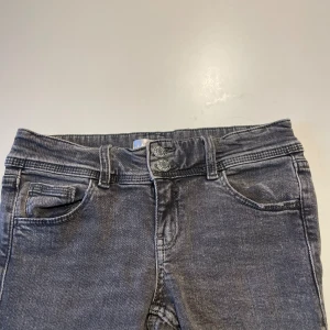 Grå bootcut jeans från Gina Tricot - Snygga grå jeans från Gina Tricot i storlek 152. Modellen har bootcut-ben och två knappar i midjan för extra cool detalj. Klassisk femficksdesign och tillverkade i mjukt jeanstyg som sitter skönt hela dagen.