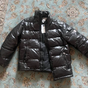 Svart pufferjacka från Moncler - Säljer en svart pufferjacka från Moncler med glansig finish och klassisk logga på ärmen. Jackan har hög krage, dragkedja framtill och två fickor med dragkedja. Perfekt för kalla dagar och riktigt snygg streetstil.