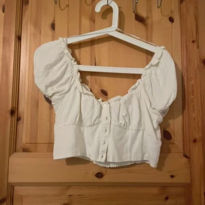 Vit croppad blus från H&M - Säljer en söt vit croppad blus från H&M med puffärmar och knappar framtill. Blusen har en rynkad kant vid halsringningen och är tillverkad i ett mjukt bomullstyg. Perfekt för en trendig och avslappnad look.