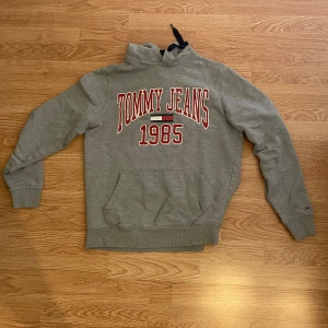 Grå hoodie Tommy Jeans 1985 - Snygg grå hoodie från Tommy Jeans med stort rött och vitt tryck 'TOMMY JEANS 1985' på bröstet. Väldigt snygg och knappt använd!