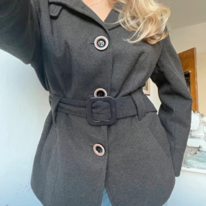 Mörkgrå kappa med bälte och stora knappar - 🖤 Mörkgrå kappa med markerat bälte  Storlek: XXL (EU 44/16) Skick: Mycket bra  Elegant svart kappa i klassisk skepparstils-modell med stora knappar och markerat bälte i midjan som framhäver figuren på ett snyggt sätt. Tyget är stadigt men mjukt, vilket gör kappan både bekväm och formfast.  Passar dig som vanligtvis bär S–M om du vill ha en oversize-look. I mycket fint skick utan tecken på användning.  Perfekt för höst och vår – tidlös, stilren och enkel att klä upp eller ner.