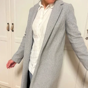 Ljusgrå kappa från Zara - Snygg ljusgrå kappa från Zara med klassisk krage och rak passform. Kappan är lång, har långa ärmar och är tillverkad i ett mjukt ullblandat material. Perfekt för dig som vill ha en stilren och tidlös look.