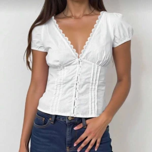 Vit blus/skjorta🤍 - Såå fin vit blus/skjorta med korta puffärmar och djup v-ringning med spetskant. Toppen har dekorativa sömmar och knappar framtill som ger en snygg siluett💕 Använd 1 gång💖