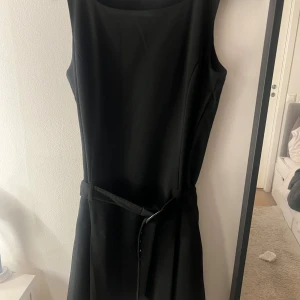Svart ärmlös klänning med bälte - Snygg svart klänning med ärmlös design och rund hals. Klänningen har ett matchande bälte i midjan som ger en stilren siluett. Perfekt för dig som gillar minimalistisk och klassisk stil. Materialet känns mjukt och har ett fint fall.