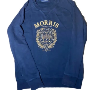 Marinblå sweatshirt från Morris - Snygg marinblå sweatshirt från Morris med stort guldfärgat tryck på bröstet med universitetsinspirerat emblem och texten 'Morris Rowing Club'. Tröjan har rund hals och långa ärmar, perfekt för en chill och sportig look.