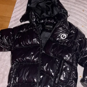  Trapstar Jacka Strlk M - Säljer en svart, glansig pufferjacka från Trapstar i storlek M. Jackan har huva, dragkedja och broderad Trapstar-logga på ryggen. Feta paneler och coolt emblem på bröstet ger jackan en streetwear-vibe. Perfekt för kalla dagar.