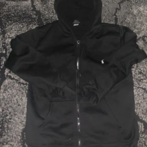 Svart hoodie från Polo Ralph Lauren - Svart hoodie med dragkedja från Polo Ralph Lauren. Klassisk design med huva, fickor framtill och den ikoniska broderade loggan i vitt på bröstet. Perfekt för chill dagar och enkel att matcha med jeans eller joggers.