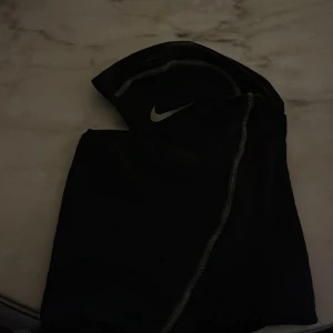  Nike ski mask - Svart balaclava från Nike med grå kontrastsömmar och klassisk Nike Swoosh-logga framtill. Tillverkad i ett stretchigt syntetmaterial som andas, perfekt för träning utomhus när det är kallt. Sitter tight runt huvudet och täcker både hals och nacke.