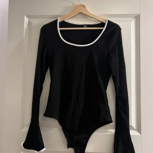 Svart långärmad bodysuit - Svart ribbad bodysuit från SHEIN med långärmade flared ärmar och vit kant vid halsringning och ärmslut. Snygg, figurnära passform och rundad hals. Helt oanvänd!
