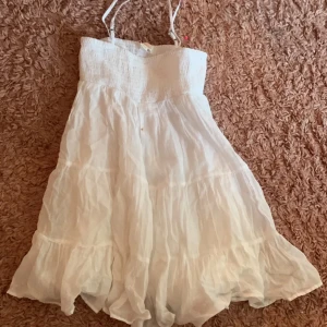 Vit sommarklänning från Gina Tricot - Supersöt vit klänning från Gina Tricot med smock upptill och tunna justerbara axelband. Kjolen är volangig och luftig, perfekt för varma dagar. Materialet är mjukt och lätt bomull, vilket gör den extra skön att bära. Passar dig som gillar en enkel och fräsch stil.