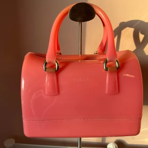 Rosa handväska från Furla i gummi - Cool rosa handväska från Furla i glansigt gummi med rundad boxig form och två rejäla handtag. Väskan har guldfärgade metalldetaljer och stängs med dragkedja. Perfekt för dig som vill sticka ut med en färgglad accessoar.