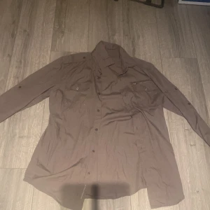 Brun skjorta från Fullcircle, storlek L - Brun skjorta från Fullcircle i storlek L, tillverkad i mjuk bomull. Skjortan har långa ärmar, knappar framtill och två bröstfickor, varav en med dragkedja. Snygg klassisk krage och detaljer som knapp på ärmen för att rulla upp. Perfekt för dig som gillar stilrena och praktiska plagg.