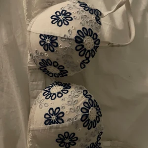 Vit blommig bh från Intimissimi - Säljer en vit bh med super push up från Intimissimi med broderade blå och vita blommor på kuporna. Axelbanden är justerbara och ryggen har spetsdetaljer. Kuporna är formade och har en mjuk känsla. Perfekt för dig som gillar unika och snygga underkläder.ena bandet är lite trasigt men den går fortfarande att använda