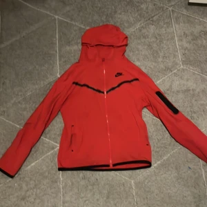 Röd Nike Tech  - Nike Tech  i rött med svarta detaljer och huva. Tröjan har hel dragkedja, två fickor fram och en svart ficka på ärmen. Snygg sportig look med svart Nike-logga på bröstet. Passar dig som gillar streetwear och sport.