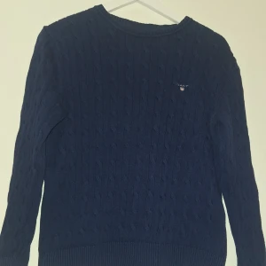 Marinblå kabelstickad tröja GANT - Kabelstickad marinblå tröja från GANT med broderad logga på bröstet. Tröjan har rund hals, ribbade muddar och lång ärm. Perfekt för dig som gillar klassisk och stilren look. Materialet är mjuk bomull som känns skönt mot huden.