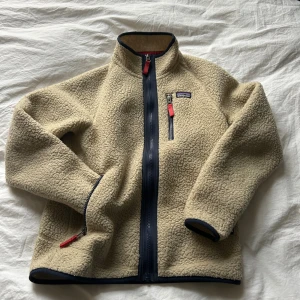 Beige teddy fleece från Patagonia L - Säljer en beige teddyjacka från Patagonia i storlek L. Jackan har mörkblå detaljer, hel dragkedja framtill och röda zip-pullers. Tillverkad i 100% återvunnen polyester, med hög krage och långärmad design. Perfekt för kalla dagar och chill stil.
