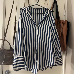 Randig blåvit skjorta från Zara - Snygg skjorta från Zara i storlek S med vertikala blå och vita ränder. Skjortan har klassisk krage, knappar framtill och långa ärmar med breda manschetter. Materialet är mjukt och lätt, perfekt för en chill och stilren look.