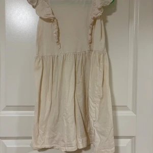 Beige kort klänning från H&M - Söt beige kort klänning från H&M med volangdetaljer vid ärmarna och lätt rynkad kjol. Klänningen har en enkel och luftig design, perfekt för varma dagar. Tillverkad i mjuk bomull för en bekväm känsla.