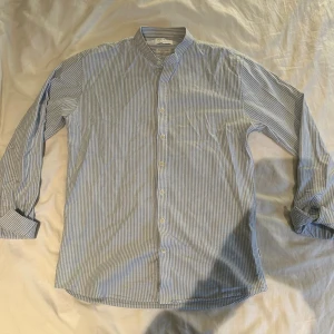 Randig skjorta från Lindbergh White - Snygg slim fit skjorta från Lindbergh White med smal ståkrage och vertikala blå och vita ränder. Skjortan har långa ärmar, knappar framtill och är gjord i mjuk bomull med superflex för extra rörelsefrihet. Perfekt för dig som gillar stilrena detaljer.