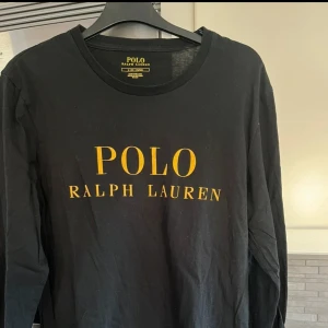 Svart långärmad tröja Polo Ralph Lauren - Svart långärmad tröja från Polo Ralph Lauren med stor gul logga framtill. Klassisk rund halsringning och mjukt bomullsmaterial. Perfekt för dig som gillar stilrena och ikoniska plagg med tydlig branding.