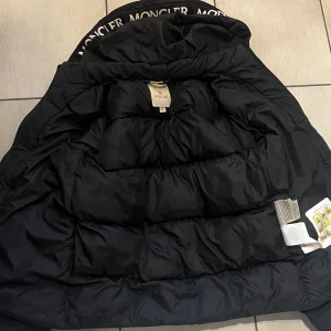 Svart Moncler dunjacka med huva - Säljer en svart dunjacka från Moncler med stor huva där Moncler-loggan är broderad i vitt. Jackan har quiltad design, dragkedja framtill och Moncler-märke på ärmen. Perfekt för kalla dagar och har en riktigt snygg, puffig look.