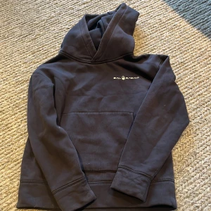 Marinblå hoodie från Sail Racing - Marinblå hoodie från Sail Racing med vit logga på bröstet. Tröjan har huva, lång ärm och en stor magficka framtill. Materialet är mjuk bomull och passformen är normal. Perfekt för chill dagar och enkel att matcha med jeans eller joggers.