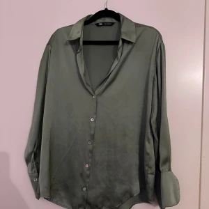 Grön satinblus från Zara - Snygg grön skjorta från Zara i mjukt satinliknande material. Klassisk krage, långärmad med breda manschetter och knappar framtill. Perfekt för dig som gillar en stilren look med lite extra glans. Passformen är loose och skjortan har en lätt skimrande finish.