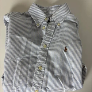 Blårandig skjorta från Ralph Lauren - Snygg blårandig skjorta från Ralph Lauren med klassisk button-down krage och broderad logga på bröstet. Custom fit-modell med vita knappar och långa ärmar. Skjortan är gjord i mjuk bomull och har en fräsch, stilren look som funkar till många outfits.