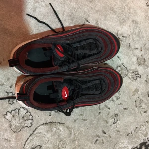 Nike Air Max 97 svart/röd sneakers - Nike Air Max 97 sneakers i svart och rött med ikoniska vågformade linjer och synlig Air-sula. Skorna har snörning, röda detaljer och en transparent sula med röd accent. Ovandelen är i syntet och textil, perfekt för dig som gillar streetstyle.