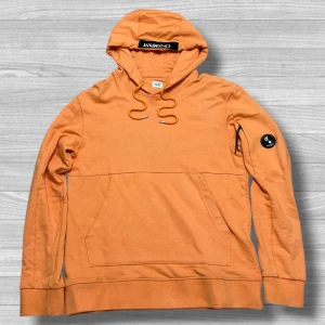 Orange hoodie från C.P. Company - Snygg orange hoodie från C.P. Company med huva och snörning. Tröjan har en klassisk känguruficka framtill och en svart patch med logga på ena ärmen. Tillverkad i mjuk bomull och har långa ärmar. Perfekt för en chill och avslappnad stil. Axlar -> Midja = 69 cm. Vänster -> Höger = 51 cm. Storlek S men passar L/M