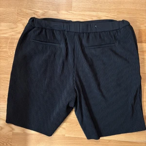 Svarta plisserade shorts  - Säljer ett par svarta shorts med plisserad struktur och relaxed fit. De har två bakfickor och är tillverkade i ett glansigt syntetmaterial som ger en modern vibe. Perfekta för dig som gillar stilrena och bekväma plagg med lite edge.