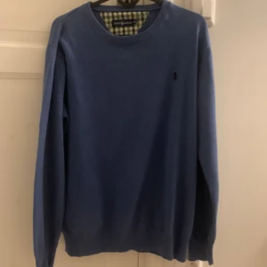 Blå stickad tröja från Ralph Lauren - Snygg blå stickad tröja från Ralph Lauren i klassisk modell med rund hals och diskret logga på bröstet. Tröjan har långa ärmar och ribbade muddar vid ärmslut och nederkant. Perfekt för dig som gillar stilrena och tidlösa plagg.