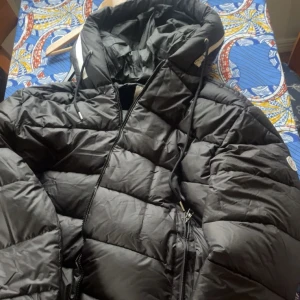 Svart Moncler pufferjacka med huva - Svart pufferjacka från Moncler med huva och snörning, klassisk logotyp på ärmen och dragkedja framtill. Jackan har quiltad design och är tillverkad i syntetmaterial med dunfyllning. Perfekt för kalla dagar och har coola detaljer som serietidningsetikett på insidan.