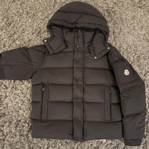 Svart Moncler pufferjacka med huva - Svart pufferjacka från Moncler med stor huva och klassisk logga på ärmen. Jackan har quiltad design, dragkedja och knappar framtill samt praktiska fickor. Perfekt för kalla dagar och har en riktigt schysst streetstil.