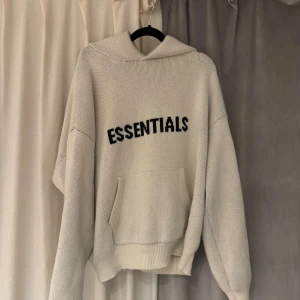 Beige Essentials hoodie med huva - Säljer en beige hoodie från Essentials med svart text framtill. Tröjan har huva, känguruficka och är tillverkad i en mjuk blandning av bomull och polyester. Perfekt för chill dagar och har en relaxed passform med långa ärmar.
