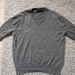 Grå v-ringad tröja Polo Ralph Lauren🙌🤩 - Snygg grå v-ringad tröja från Polo Ralph Lauren i mjuk pima bomull. Klassisk design med diskret broderad logga på bröstet och ribbade muddar vid ärmslut och nederkant. Perfekt för dig som gillar stilrena och tidlösa plagg.🙌🤩