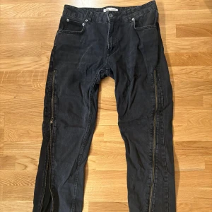 Svarta jeans med dragkedjor från ZARA - Säljer ett par svarta jeans från ZARA med coola dragkedjor längs hela utsidan av benen. Jeansen har klassisk femficksdesign och är tillverkade i jeansmaterial med en mörk tvätt. Passformen är rak och detaljerna ger en edgy vibe.