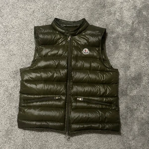 Moncler gui - Snygg mörkgrön dunväst från Moncler med quiltad design. Västen har två praktiska fickor med dragkedja och klassisk Moncler-logga på bröstet. Perfekt att slänga över en hoodie för extra värme. Stängs med dragkedja framtill. Det är storlek 2 så det är S/M. Hör av er vid minsta lilla fråga och priset är ej hugget i sten