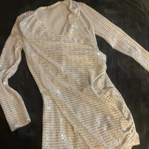 Glittrig vit kortklänning från NLY One - Supersnygg vit kortklänning från NLY One med glittriga paljetter och diskret randigt mönster. Klänningen har omlott-design, v-ringning och långa ärmar. 