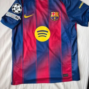 FC Barcelona Raphinha 11 matchtröja - Säljer en officiell FC Barcelona fotbollströja med Raphinha 11-tryck. Tröjan är kortärmad, har klassiska blå och röda ränder, gula detaljer och klubbmärke på bröstet. Tillverkad i lätt och ventilerande material med Champions League-märke på ärmen.