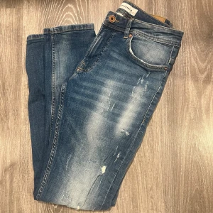 Blå slitna jeans  - Snygga blå jeans med slitningar och tvättade detaljer på framsidan. Modellen har klassisk femficksdesign, knapp och dragkedja i midjan samt smal passform. Perfekt för dig som gillar en avslappnad och trendig look.