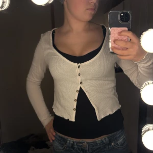 Vit croppad ribbad blus från H&M - Säljer en vit croppad blus från H&M med ribbad struktur och v-ringning. Toppen har långa ärmar, knappar framtill och en vågig kant vid nederdelen. Perfekt för dig som gillar en chill och trendig stil.