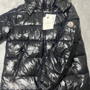 Svart Moncler pufferjacka M - Svart pufferjacka från Moncler med glansig finish, ikonisk logga på ärmen och dragkedjor på fickorna. Jackan har hög krage och är fylld med vitt gåsdun för extra värme. Perfekt för dig som vill ha en snygg och lyxig vinterjacka.