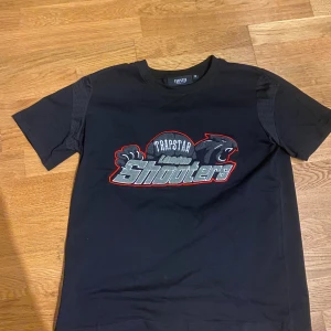 Trapstar svart t-shirt London Shooters - Svart t-shirt från Trapstar med fet broderad London Shooters-logga i grått, vitt och rött på bröstet. Den passar som en small. Rund hals och korta ärmar. Materialet är 80% bomull och 20% polyester, vilket gör den både mjuk och slitstark. Perfekt för dig som gillar streetwear och snygga detaljer.