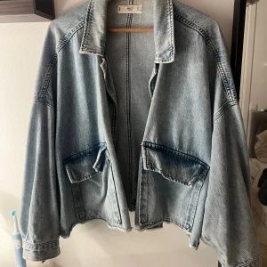 Oversized jeansjacka från MNG Denim - Säljer en ljusblå oversized jeansjacka från MNG Denim. Jackan har stora fickor framtill, klassisk krage och avslappnad passform. Perfekt för dig som gillar en chill och trendig look. Materialet är 100% denim och jackan har en rå, lite sliten kant nertill.