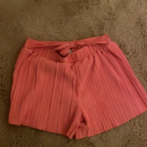 Plisserade rosa shorts från H&M - Säljer ett par snygga plisserade shorts från H&M i en härlig rosa färg. Shortsen har hög midja och ett brett knytband framtill som ger en extra cool touch. Perfekta för varma sommardagar och riktigt sköna att ha på sig.