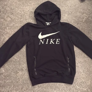 Svart Nike hoodie med dragkedjor - Svart hoodie från Nike med stor vit logga på bröstet. Tröjan har huva med snörning och två praktiska fickor med dragkedja. Långärmad och tillverkad i mjukt syntetmaterial, perfekt för en sportig och avslappnad stil.