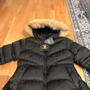 Svart pufferjacka från Zavetti Canada - Jet black pufferjacka från Zavetti Canada med fuskpäls på huvan. Kort passform och sitter fint mot kroppen. Aldrig använd, kommer med prislapp. Köpte en M men den passar mer som en S så ha det i åtanke 