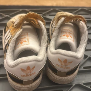Adidas Superstar med zebramönster - Unika Adidas Superstar sneakers med zebramönster i svart, beige och brunt. Skorna har klassiska vita ränder på sidorna, vit tå och häl med orange Adidas-logga. Ovandelen är i mocka och skinn, med kardborreknäppning för enkel påtagning.
