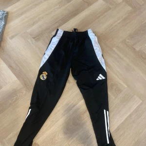 Svarta Adidas Real Madrid träningsbyxor - Svarta träningsbyxor från Adidas med Real Madrid-logga på benet och vita detaljer längs sidorna och benen. Byxorna har snörning i midjan och är tillverkade i ett syntetmaterial som passar perfekt för sport och träning.